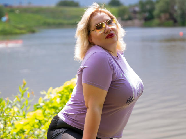 AngelinaGlam with blonde hair, wearing a tight purple shirt showing her big tits, black pants, and gold sunglasses, leaning forward by a sunny river.