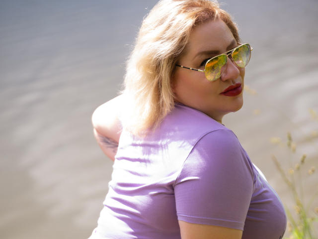 AngelinaGlam with blonde hair, wearing green reflective sunglasses, a tight purple shirt, and red lipstick, looking back over her shoulder, showing her small tits. Water background.