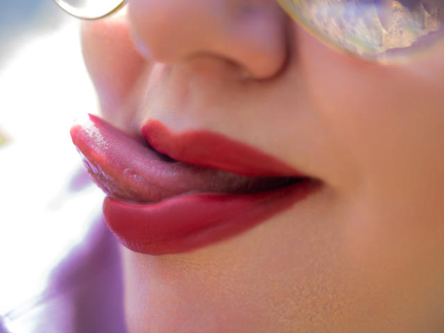 AngelinaGlam's pink tongue is out, licking her red lipstick-covered bottom lip. Her smooth, fair skin and shiny nose are visible. She's wearing a reflective gold ring.