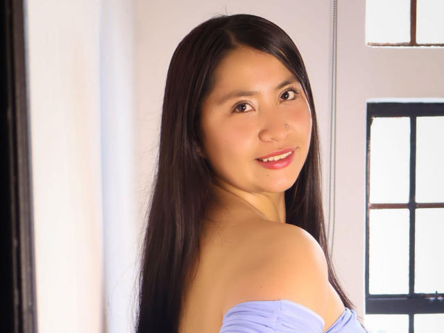 AshleyWalkerI with long black hair, smiling, light skin, small nose ring, wearing a purple off-shoulder top, looking over shoulder, soft light, window in background.
