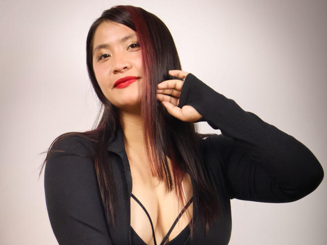 AshleyWalkerI has red lipstick, straight black hair with a red streak, and wears a black cardigan with a low-cut black top showing her tits. She's smiling, touching her hair.