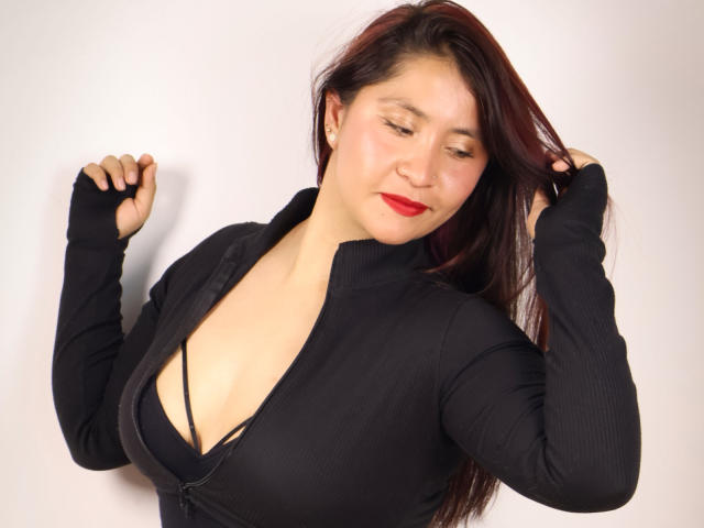 AshleyWalkerI with red lipstick, black zip-up top, and visible cleavage, holding her hair with hands, looking down, smiling slightly, in a studio shot, wearing black fingerless gloves.