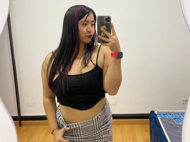 AshleyWalkerI with long black hair, red lipstick, black crop top showing big tits, and checkered pants, taking a mirror selfie, wearing a red watch, in a store.