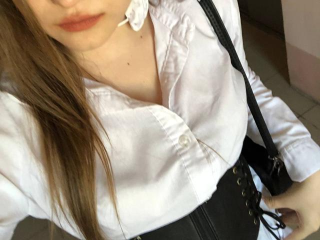 Bubochka has long brown hair, a white button-up shirt, and a black corset with lace-up detail. Her lips are slightly parted with a small white rose in her mouth.