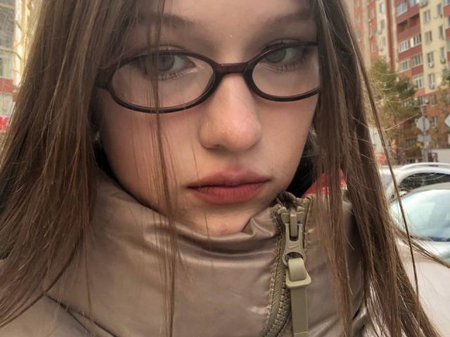Bubochka with brown glasses, pale skin, and long brown hair, wearing a beige puffy jacket, has her lips slightly parted, looking directly at the camera with a serious expression.
