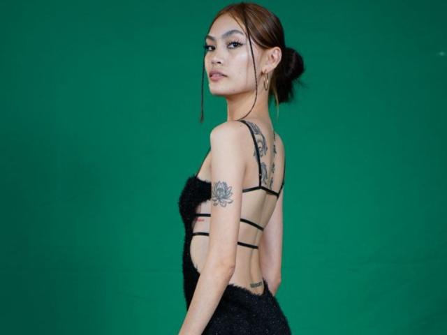 XAkariMorisX in a black strapless fur dress, showing tattoos on her back and arm, looking over her shoulder with brown hair in a bun, green background.