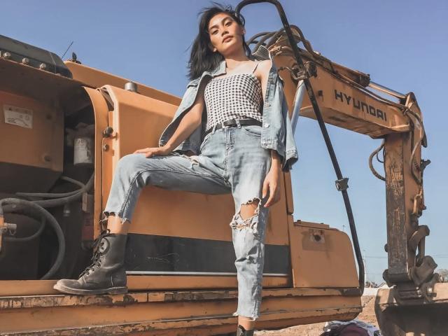 XAkariMorisX standing with one boot on a yellow Hymachi excavator, wearing a black and white checkered top, ripped jeans, and black boots, with black hair and a confident look.