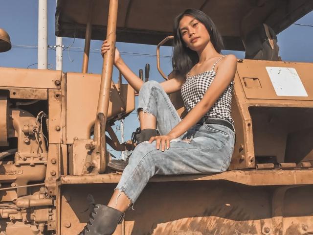 XAkariMorisX sits on a rusty, brown bulldozer, wearing a black and white checkered top, blue ripped jeans, and black boots, with her hand on the control stick.