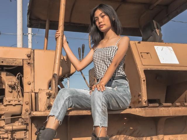 XAkariMorisX sitting on a dirty construction machine, wearing a black-and-white checkered top, ripped blue jeans, and black boots, holding a wooden pole, with medium tits and dark hair.