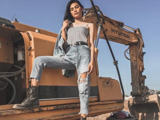 XAkariMorisX stands in front of a yellow HYDROCAT excavator, wearing a black-and-white strapless top, ripped blue jeans, and black boots, with her left foot on the machine.