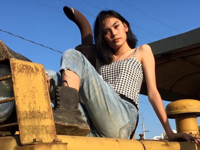 XAkariMorisX with black hair, wearing a checkered top and ripped jeans, sits on rusty yellow machinery, black boots, looking down, blue sky in background.