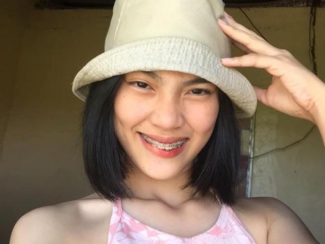 XAkariMorisX with black shoulder-length hair, wearing a beige bucket hat, smiling with metal braces, and pink patterned tank top, holding hat with right hand.