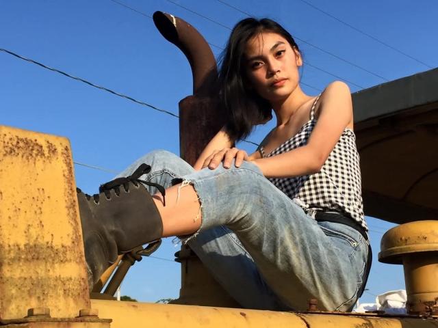 XAkariMorisX sits on a rusty yellow metal structure, wearing a black-and-white checkered tank top, ripped blue jeans, and black boots, with a blue sky background.