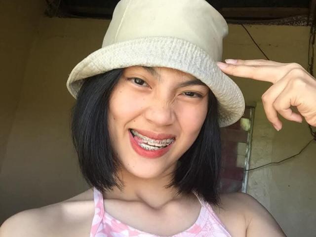 XAkariMorisX with black shoulder-length hair, smiling with metal braces, wearing a beige bucket hat, and a pink tie-dye halter top, pointing at the hat with her right hand.