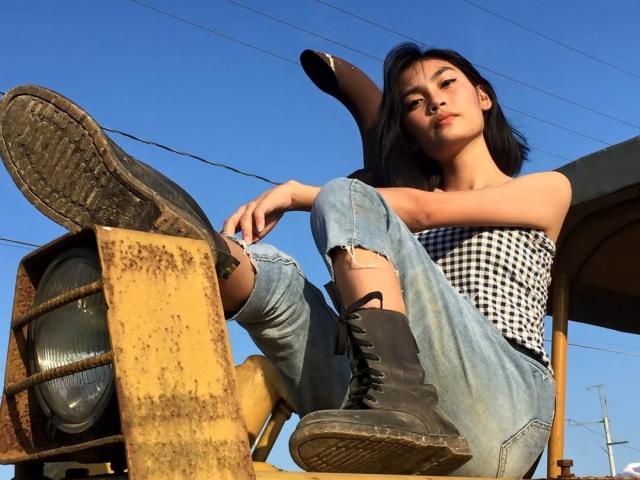 XAkariMorisX with black hair, wearing a black-and-white checkered top, ripped jeans, and black boots, sitting on a rusty yellow machine, looking down with a relaxed expression.