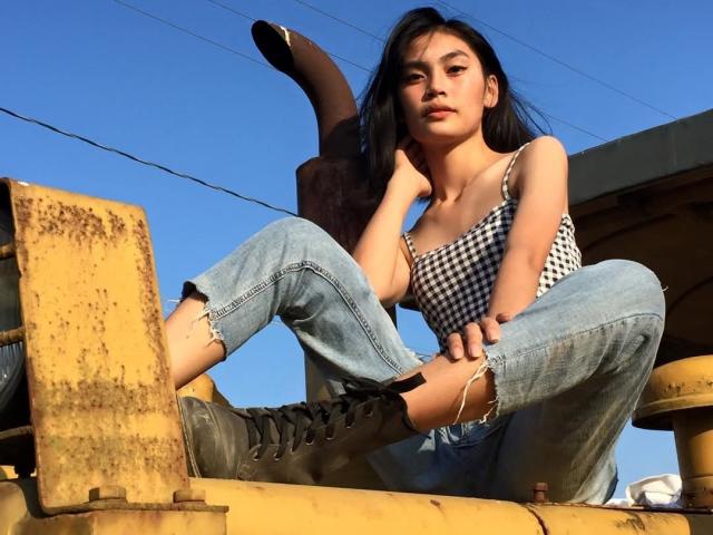 XAkariMorisX with long black hair, checkered top, and ripped jeans, sitting on a rusty yellow machine, wearing black boots, looking down with a slightly bored expression.