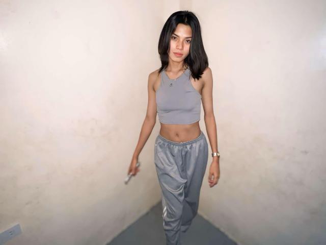 XHellyMariaX met zwart haar dat tot op de schouders komt, een grijze sportbeha, waarbij kleine borsten zichtbaar zijn, en grijze satijnpantalon, staat in een hoek, houdt een sigaret vast en draagt een gouden horloge..