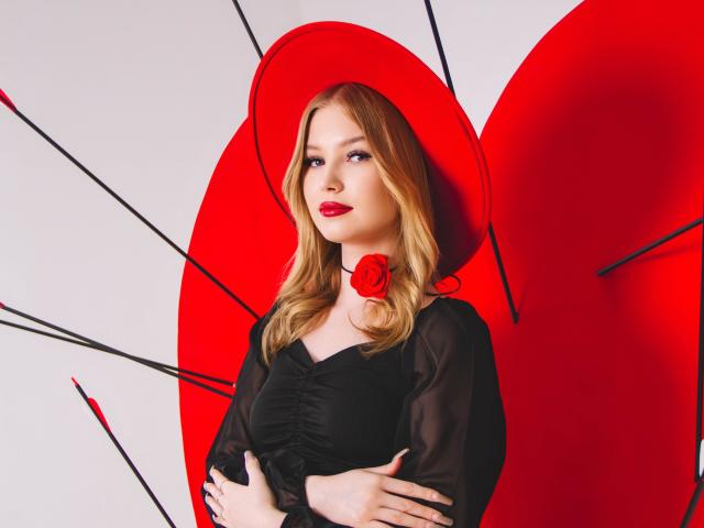 NicoleBrian with blonde hair, red lipstick, black sheer dress, red rose choker, red hat, arms crossed, black umbrella handles in background. She's a sexy, pretty, cute, attractive woman.