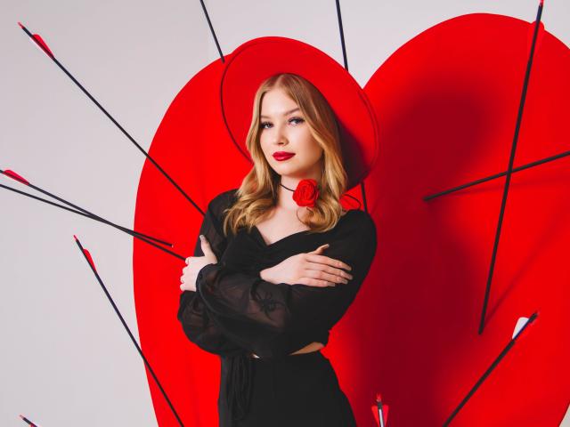 NicoleBrian with blonde hair, red hat, black sheer top, red rose, and crossed arms, standing against a large red heart with black arrows. Her tits are small.