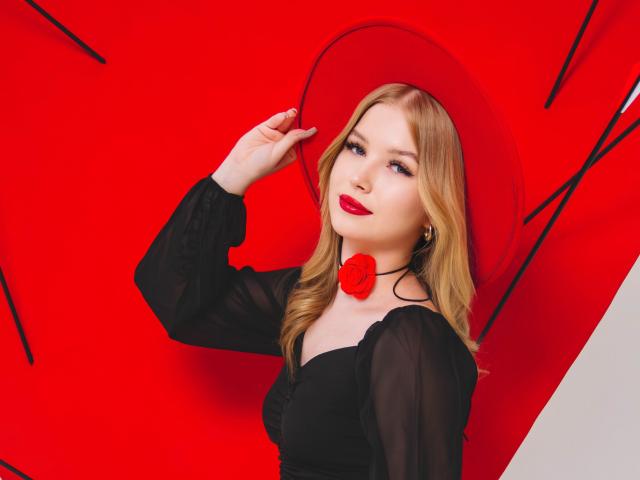 NicoleBrian with blonde hair, wearing a black sheer dress, red rose choker, and matching red hat, has red lipstick and blue eyes, touching her hat with a confident look.
