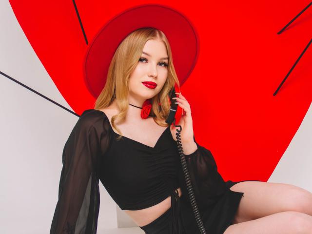 NicoleBrian with blonde hair, red hat, black sheer dress, holding a phone, red lips, black choker, sitting, showing her tits, legs crossed, white background, red geometric pattern.