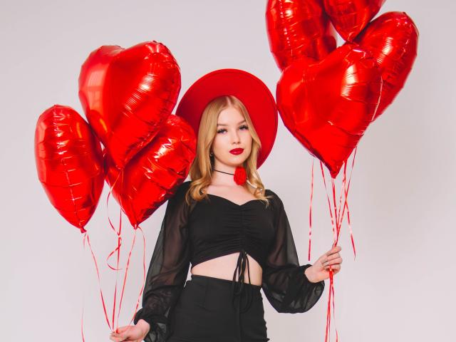 NicoleBrian with red heart balloons, wearing a black sheer top, black skirt, and red hat, holding red heart balloons, red lipstick, blonde hair, white background.