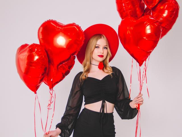 NicoleBrian with red heart balloons, wearing a black sheer top, black pants, red hat, holding three shiny red heart balloons, has blonde hair, red lipstick, and a cute face.