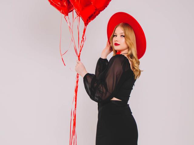 NicoleBrian with blonde hair, red hat, black sheer dress, holding red balloons, showing ass, red lipstick, looking back, right hand on hat.