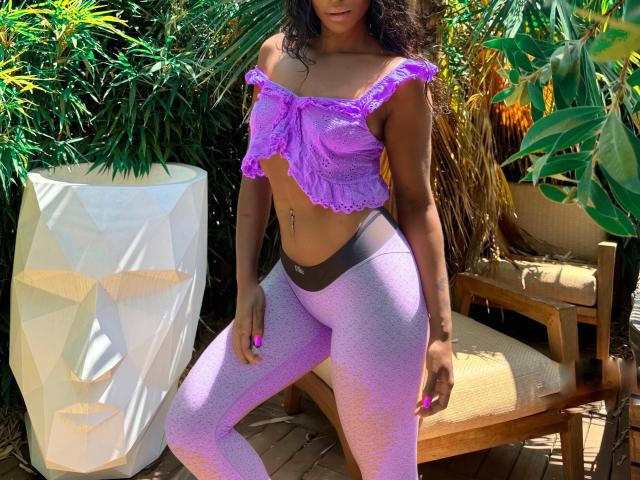 Ciarana with purple off-shoulder top, showing small tits, wearing purple glittery leggings, standing in sunlit garden, curly dark hair, tan skin, navel piercing, wooden chair in background.