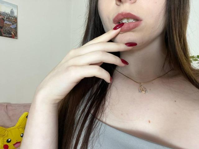Valissia, avec des ongles rouges, un doigt sur ses lèvres, une peau claire, des cheveux bruns, un petit collier en or, des lèvres rosées légèrement entrouvertes, un haut blanc à épaule dénudée, un oreiller Pikachu en arrière-plan..