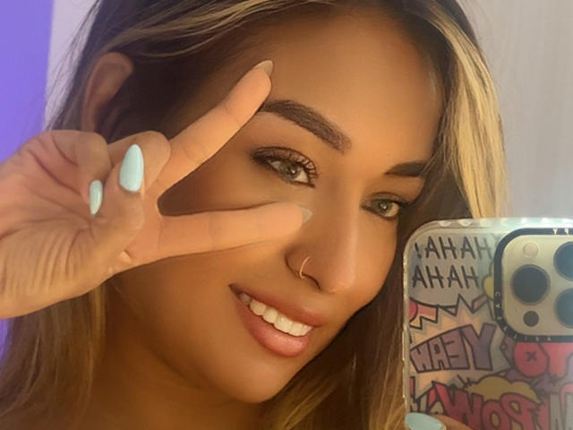 Diana Hopson, con piel morena, ojos marrones y reflejos rubios, haciendo el gesto de la paz con su mano izquierda, sonriendo, mostrando su piercing rosa en la nariz y sus uñas azul claro..