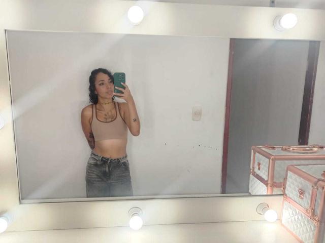 SilvanaXOwen with dark curly hair, tan bra, and high-waisted jeans, standing in front of a mirror, holding a phone, showing a tattoo on her right arm.