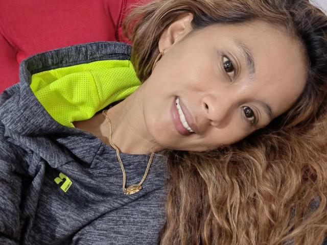 AkeshaStar, con cabello castaño ondulado, sonriendo, vestida con una sudadera gris con forro verde neón, un collar dorado y pequeños aretes, acostada sobre una superficie roja..