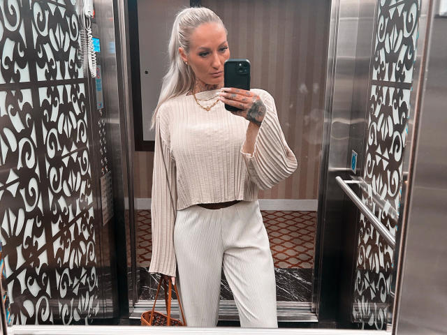 Shiva69-hot with long silver hair, white ribbed long-sleeve top, white high-waisted pants, holding phone, mirror selfie, small tattoo on left wrist, brown purse hanging.
