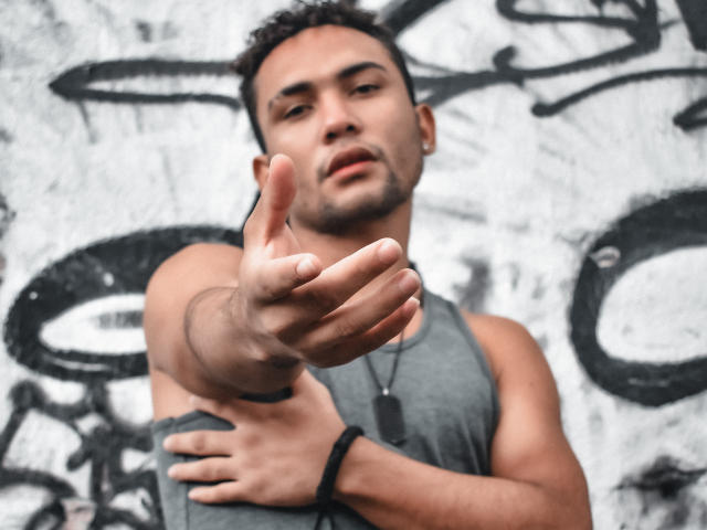 DjesusBlaise mit kurzen, lockigen Haaren, gebräunter Haut und einem grauen Tanktop, der nach vorne zeigt und einen ernsten Gesichtsausdruck hat, trägt eine Hundemarkenkette und ein schwarzes Armband..