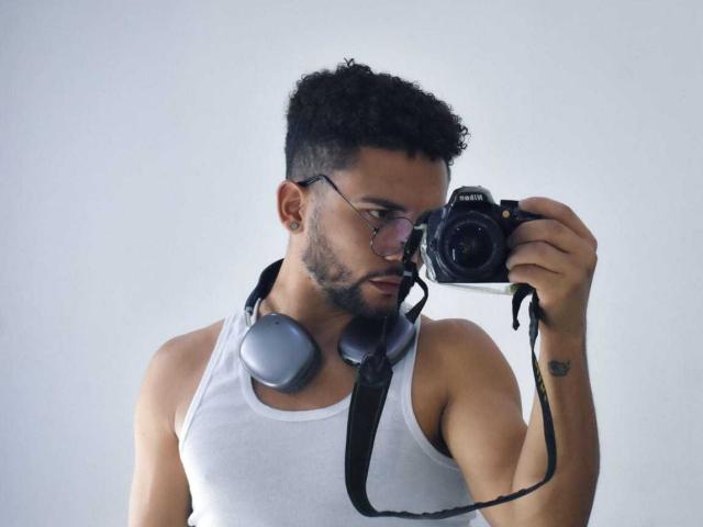 DjesusBlaise, avec des cheveux noirs bouclés, des lunettes, un débardeur blanc, tenant un appareil photo Canon, des écouteurs gris autour du cou, une expression concentrée, un tatouage au poignet droit..