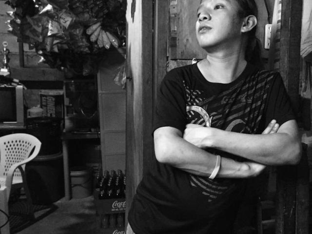 Black-and-white photo of NaughtySexFrYou, a slim Asian woman with small tits, arms crossed, looking up, wearing a dark T-shirt, standing next to a wooden wall.