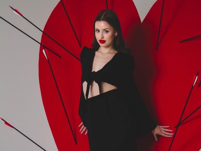CherylFoxy with red lipstick, wearing a black, cut-out, long-sleeve top, showing her tits, standing in front of a large red heart with black arrows.