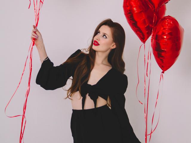 CherylFoxy with long brown hair, red lipstick, black top, holding red heart balloons, her tits slightly visible, white background.