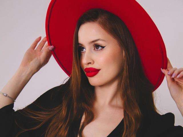 CherylFoxy with red hat, holding it, showing off bright red lipstick, long brown hair, black top, white background, pink nails, silver bracelet, and a slight smile.