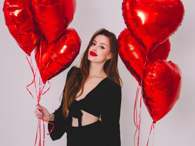 CherylFoxy with red heart-shaped balloons, wearing a black cut-out dress, holding them, has red lipstick, long brown hair, and a cute, sexy look.