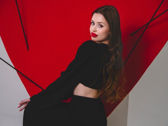 CherylFoxy with long brown hair, red lipstick, black crop top, and black pants, looking over her shoulder, standing against a red triangle with black lines.