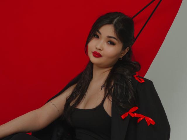 TeyaFaber with black hair, red lipstick, black tank top, and black jacket with red ribbons, sitting, showing small tits, confident smile, light skin, subtle tattoo on arm.