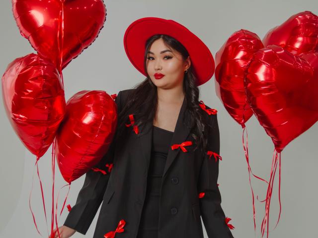 TeyaFaber with red heart balloons, wearing black coat with red bows, red hat, and red lipstick, holding four shiny red heart balloons. Her tits are slightly visible under coat.