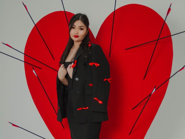 TeyaFaber stands in front of a red heart with arrows, wearing a black suit with red bow patterns, holding her collar, with long black hair and red lipstick.