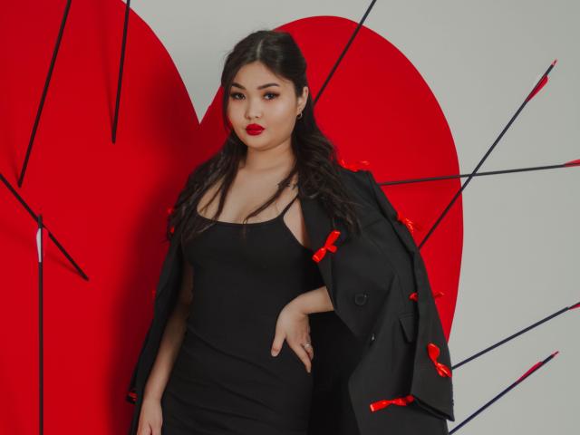 TeyaFaber with dark hair, red lipstick, wearing black dress, black jacket with red bows, standing, arm resting on hip, looking at camera, medium shot, sexy, curvy, cute.