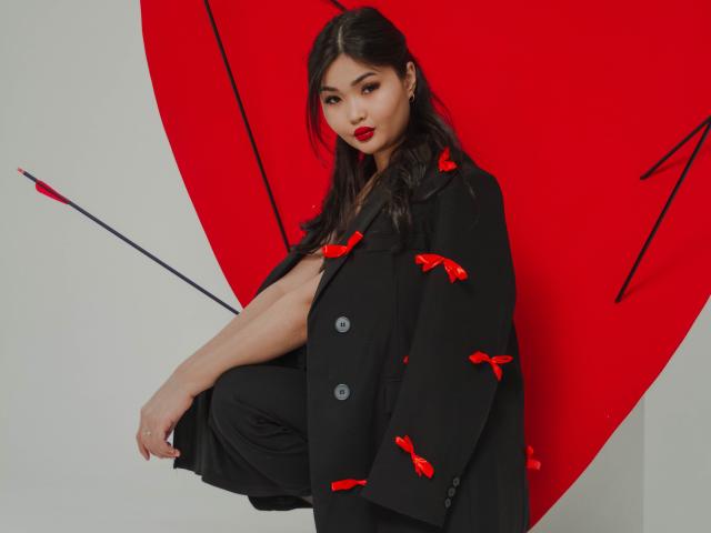 TeyaFaber with dark hair, red lipstick, black coat with red bows, black pants, holding a red umbrella with black tips, standing against a red heart-shaped background.