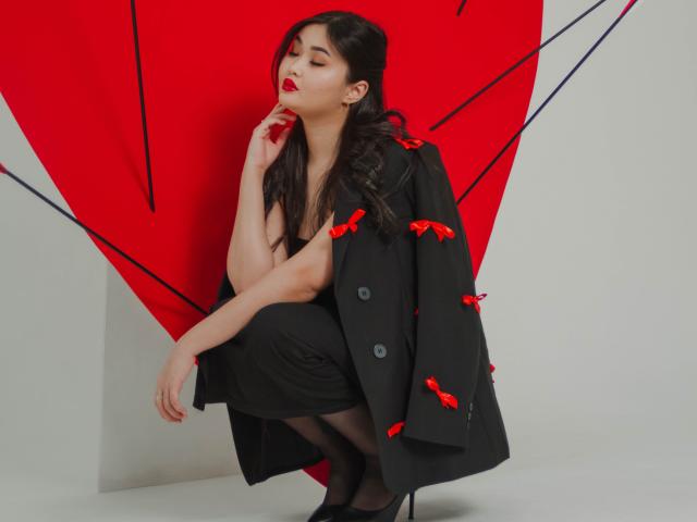 TeyaFaber with dark hair, red lipstick, crouching, wearing black coat with red bows, black dress, black stockings, black heels, thinking with finger on lip, sexy, confident.