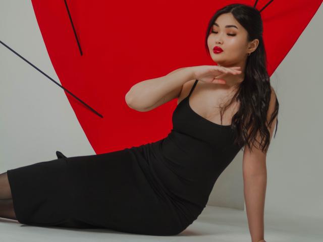 TeyaFaber with dark hair, red lipstick, black dress, and sheer black stockings, sitting, arm raised, eyes closed, showing off her tits and ass. Red triangular background.
