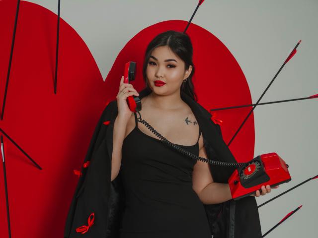 TeyaFaber with red lipstick, black dress, holding two red landline phones, black cape with red ribbon, small star tattoo on right shoulder, dark hair, white background.